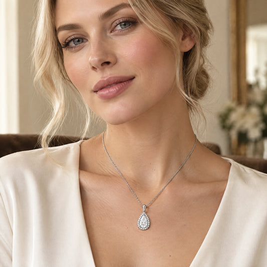 A woman wearing a silver teardrop pendant necklace with a halo of small clear stones, paired with a cream satin top.