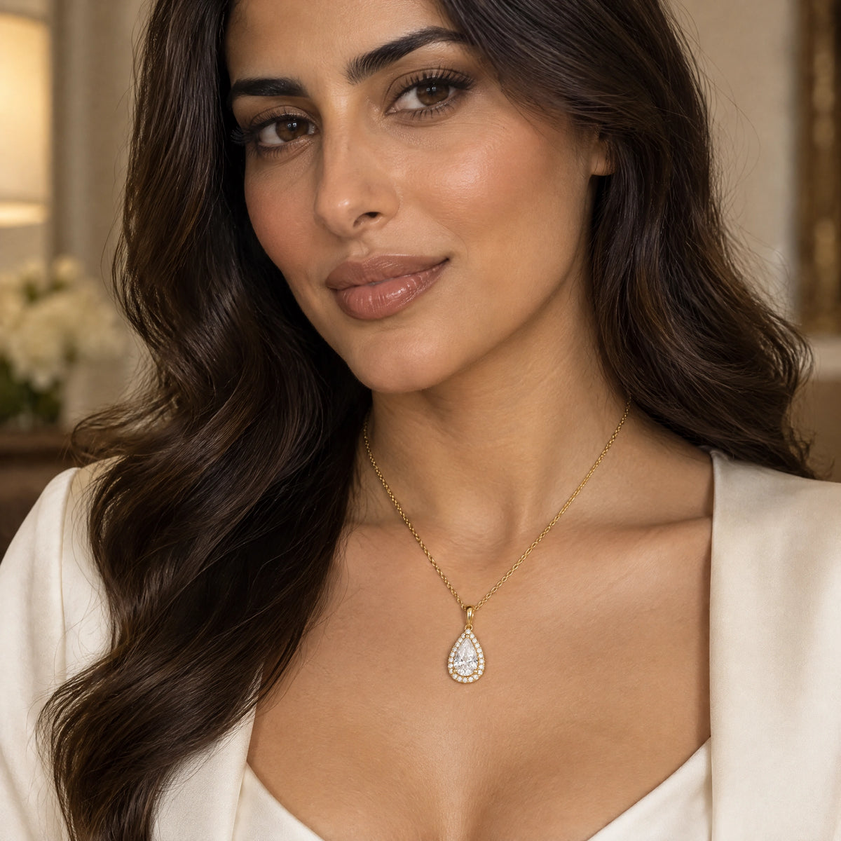 A woman with long wavy dark brown hair wears a delicate gold chain necklace with a teardrop-shaped clear gemstone pendant, styled with a white satin top in a softly lit indoor setting.