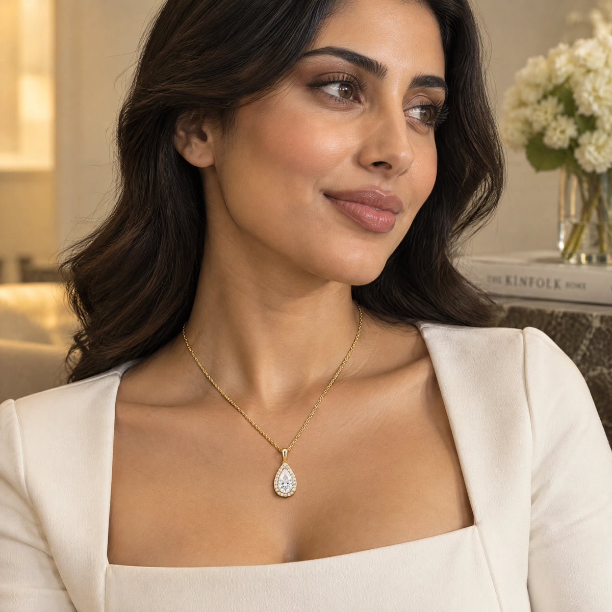 A woman wearing a delicate gold chain necklace with a teardrop-shaped pendant set with clear stones, styled with a white top in a softly lit indoor setting.