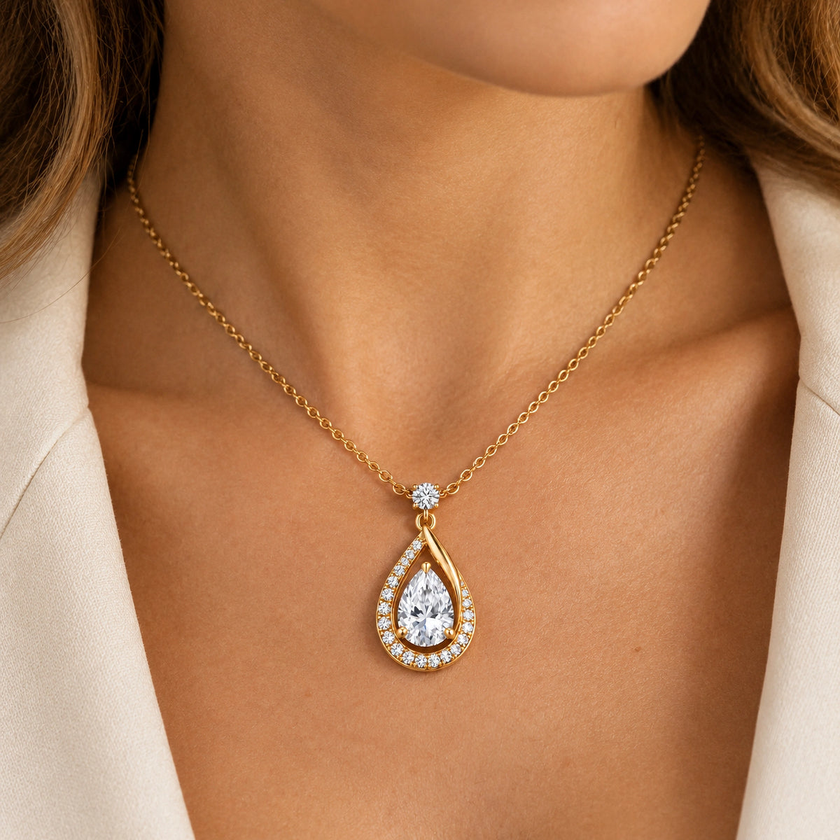 A gold teardrop pendant necklace with a clear teardrop gemstone and a halo of small clear stones, worn on a model's neck.