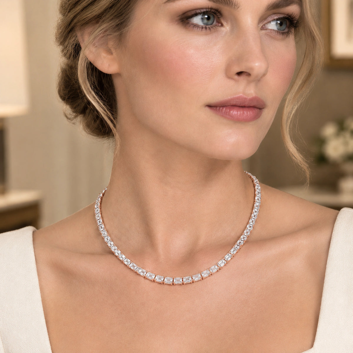 A close-up of a woman wearing a delicate tennis-style necklace with clear gemstones in a soft warm-toned indoor setting.